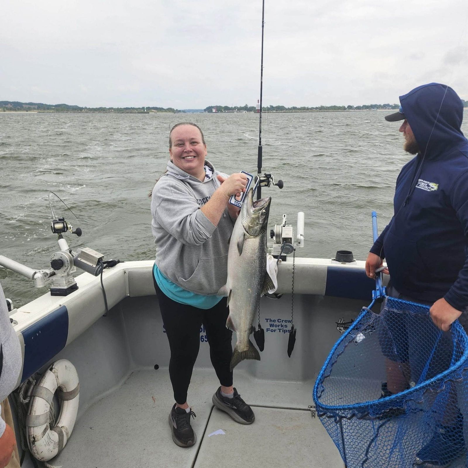 A smiling angler holds up a freshly caught fish on deck with a crew member and net nearby