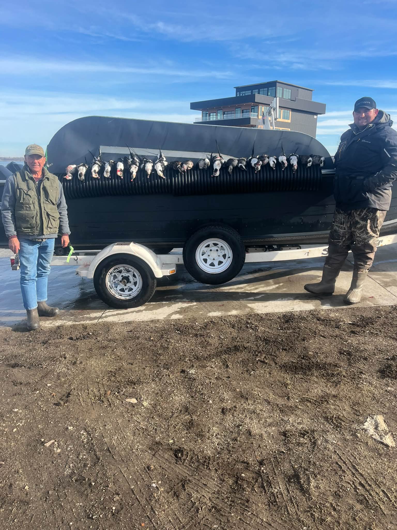Two anglers display a successful catch of numerous fish lined up in a black tank trailer at the dock, with a modern charter building visible in the background.