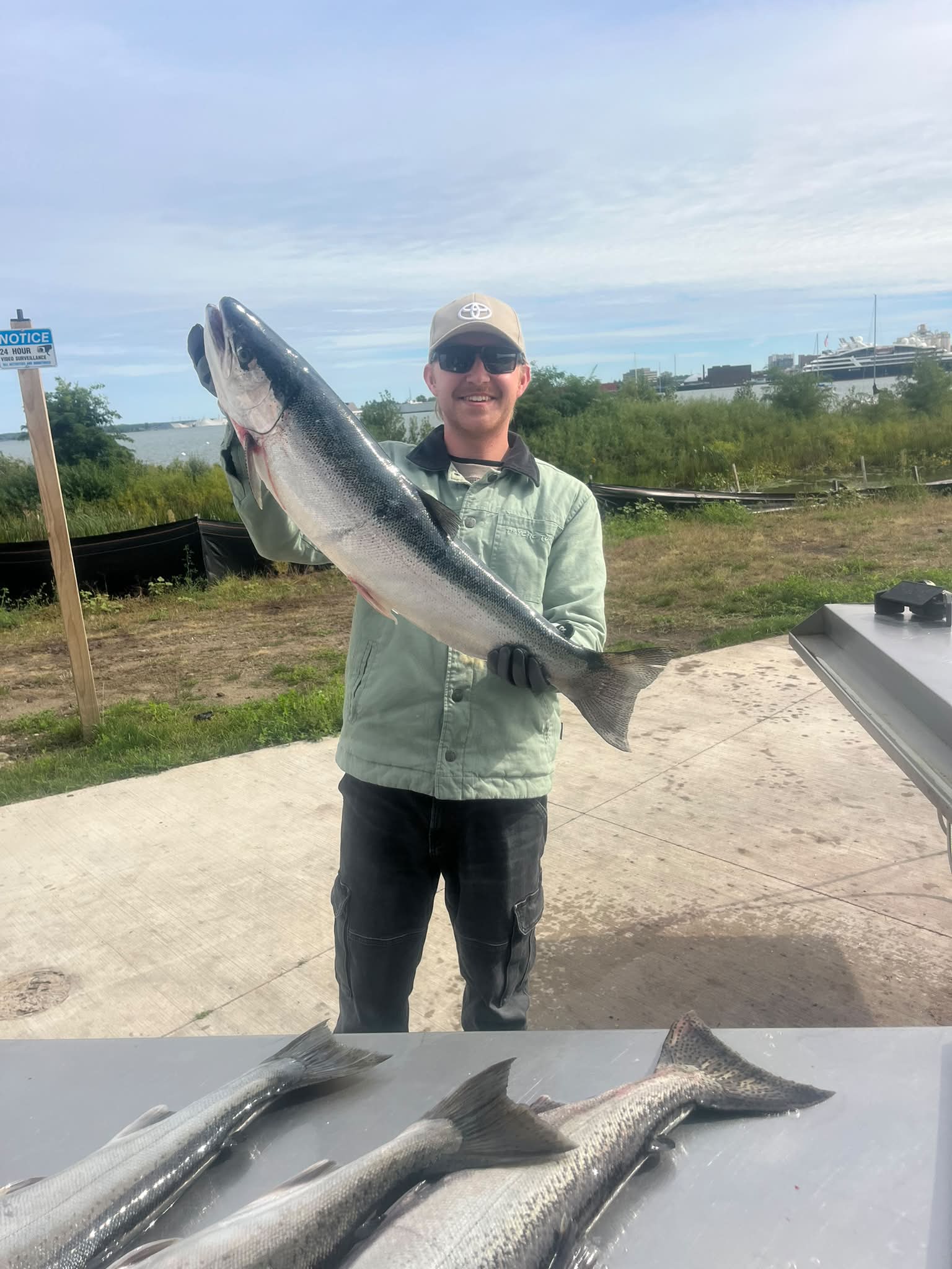 A smiling angler holds up a large silver salmon catch at the dock, with additional fish displayed on the table below.