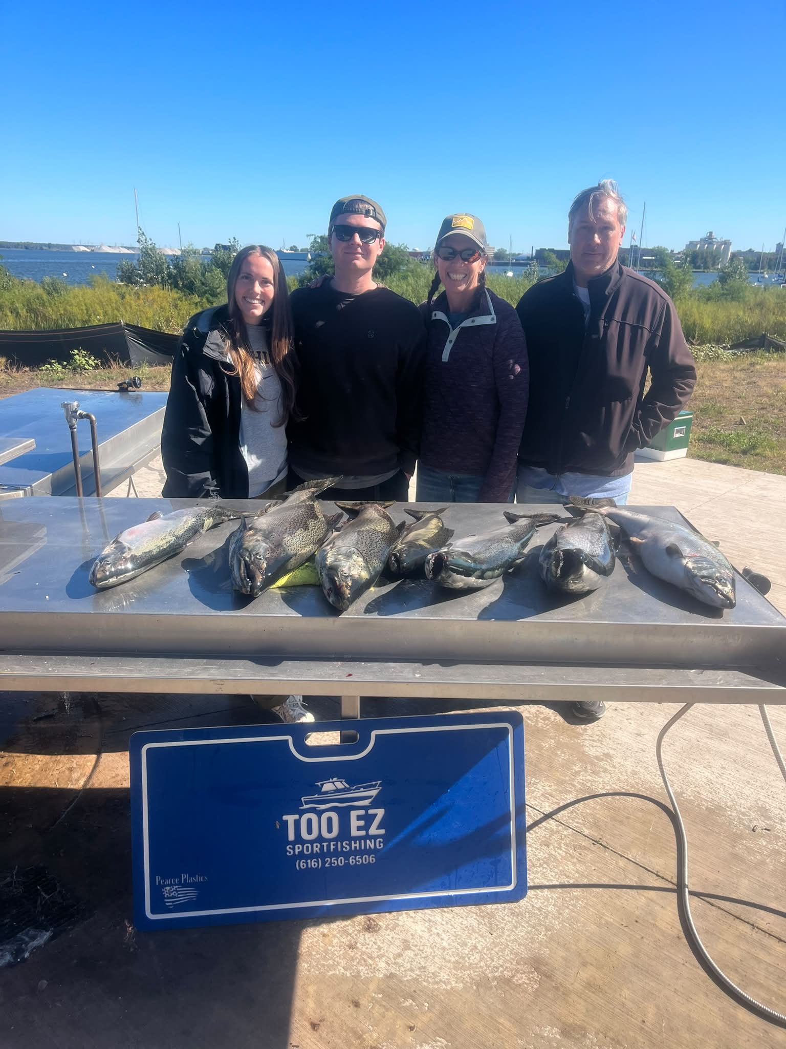 Four anglers pose with their catch of multiple fish on a table at Too Ez Sportfishing's waterside location with marina visible in the background.